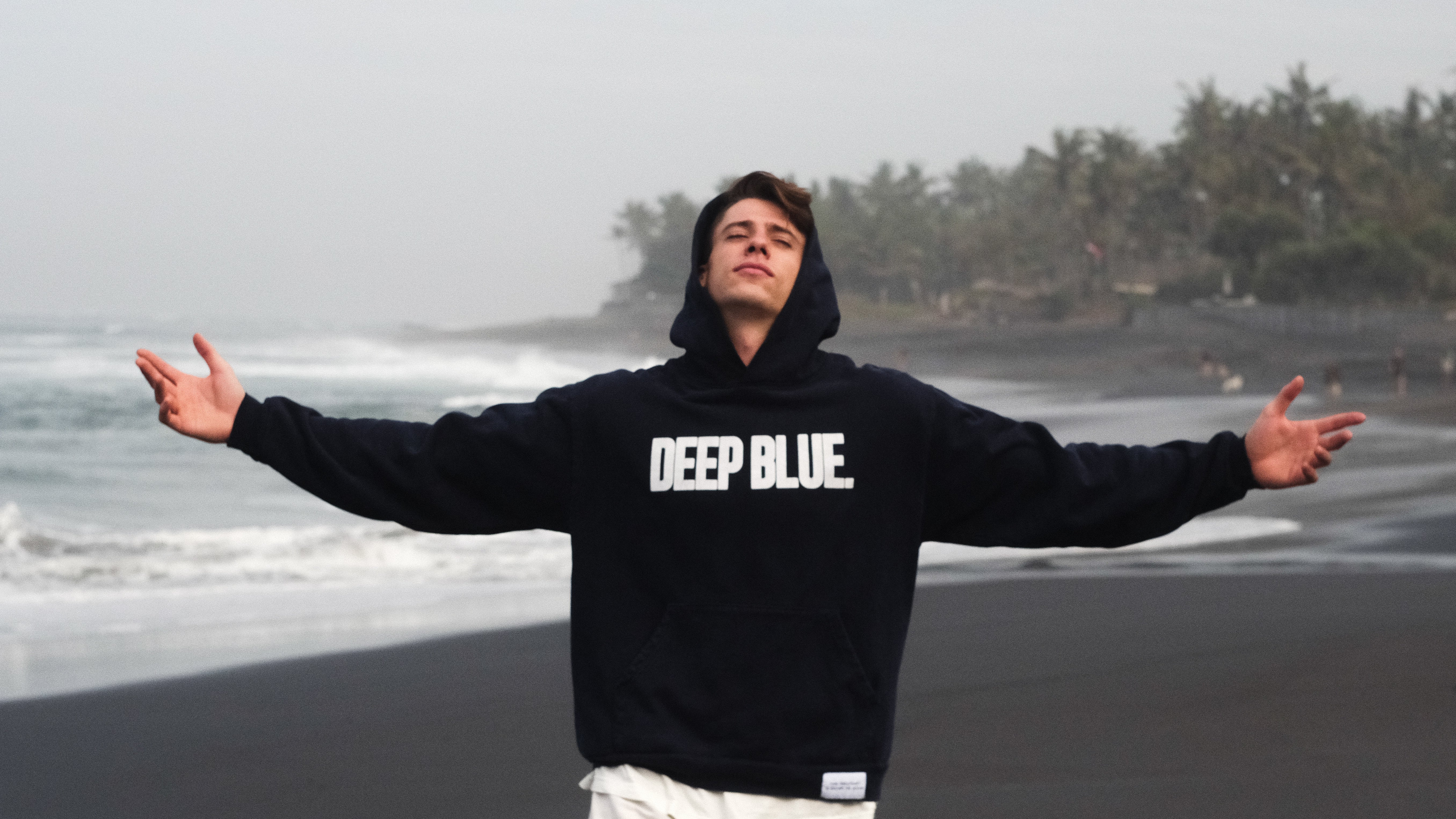 Person wearing a black hoodie with 'DEEP BLUE' text on a beach